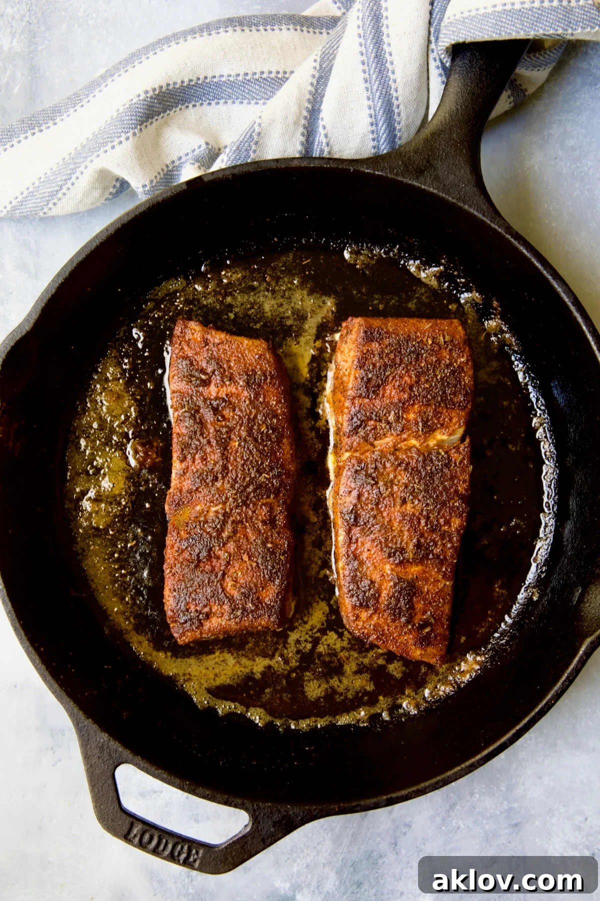 Two pieces of halibut coated with blackening seasoning in cast iron skillet after being cooked with blue striped towel wrapped around the handle.