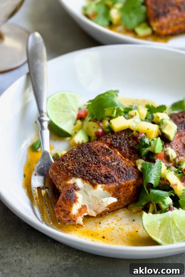Blackened Halibut with Mango - Avocado Salsa on white plate with fork garnished with cilantro and lime wedge with a piece removed to show the inside of the fish.