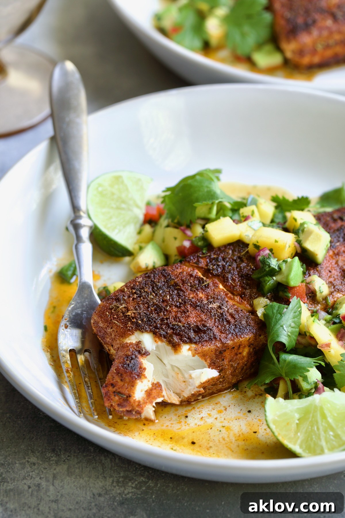 Blackened Halibut with Mango - Avocado Salsa on white plate with fork garnished with cilantro and lime wedge with a piece removed to show the inside of the fish.