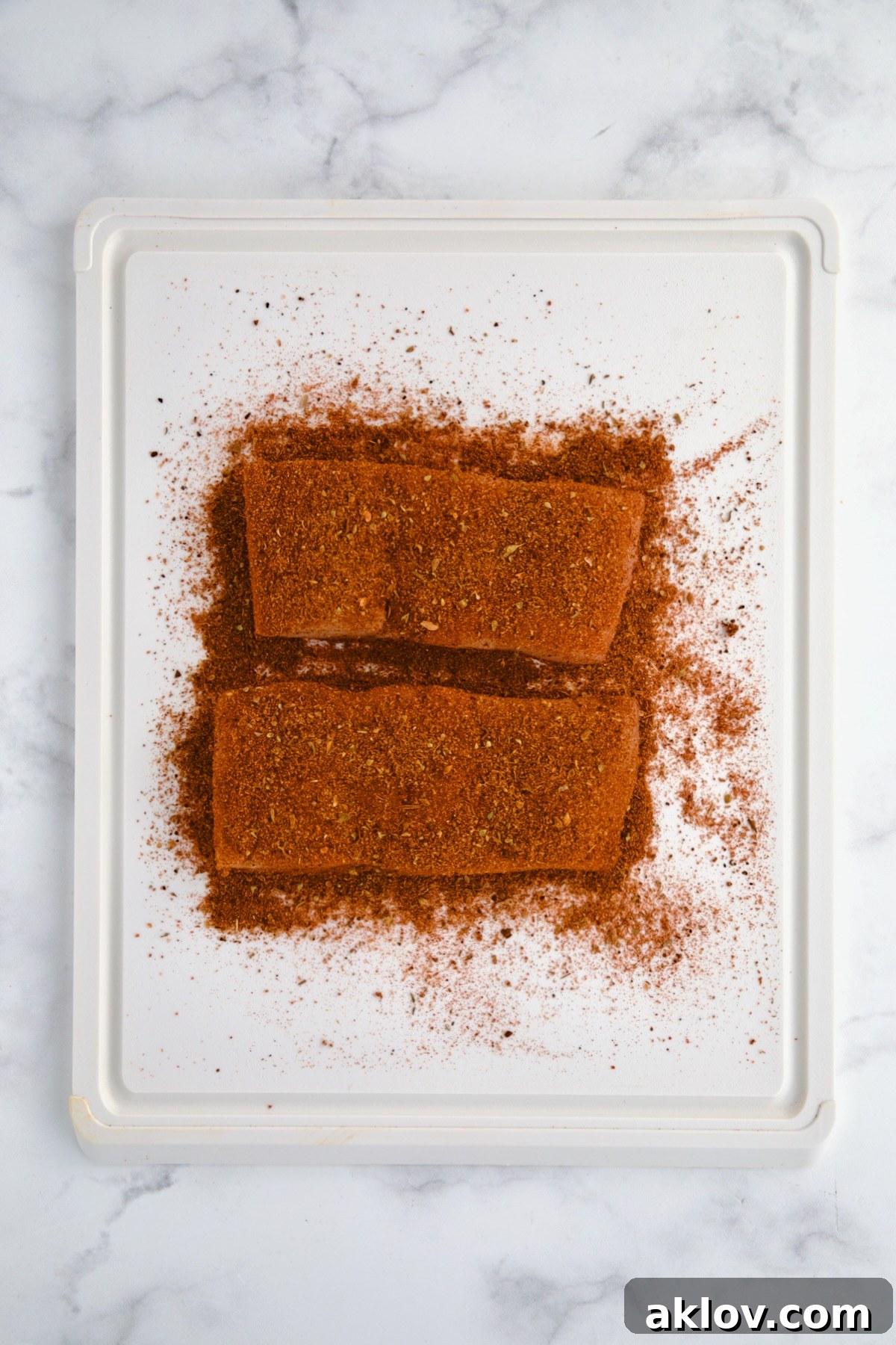 Two pieces of uncooked halibut coated with blackening seasoning on white plastic cutting board.