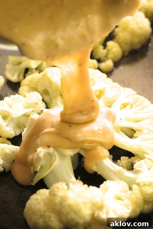 Thick, creamy cheese sauce being poured over roasted cauliflower steaks on a baking sheet.
