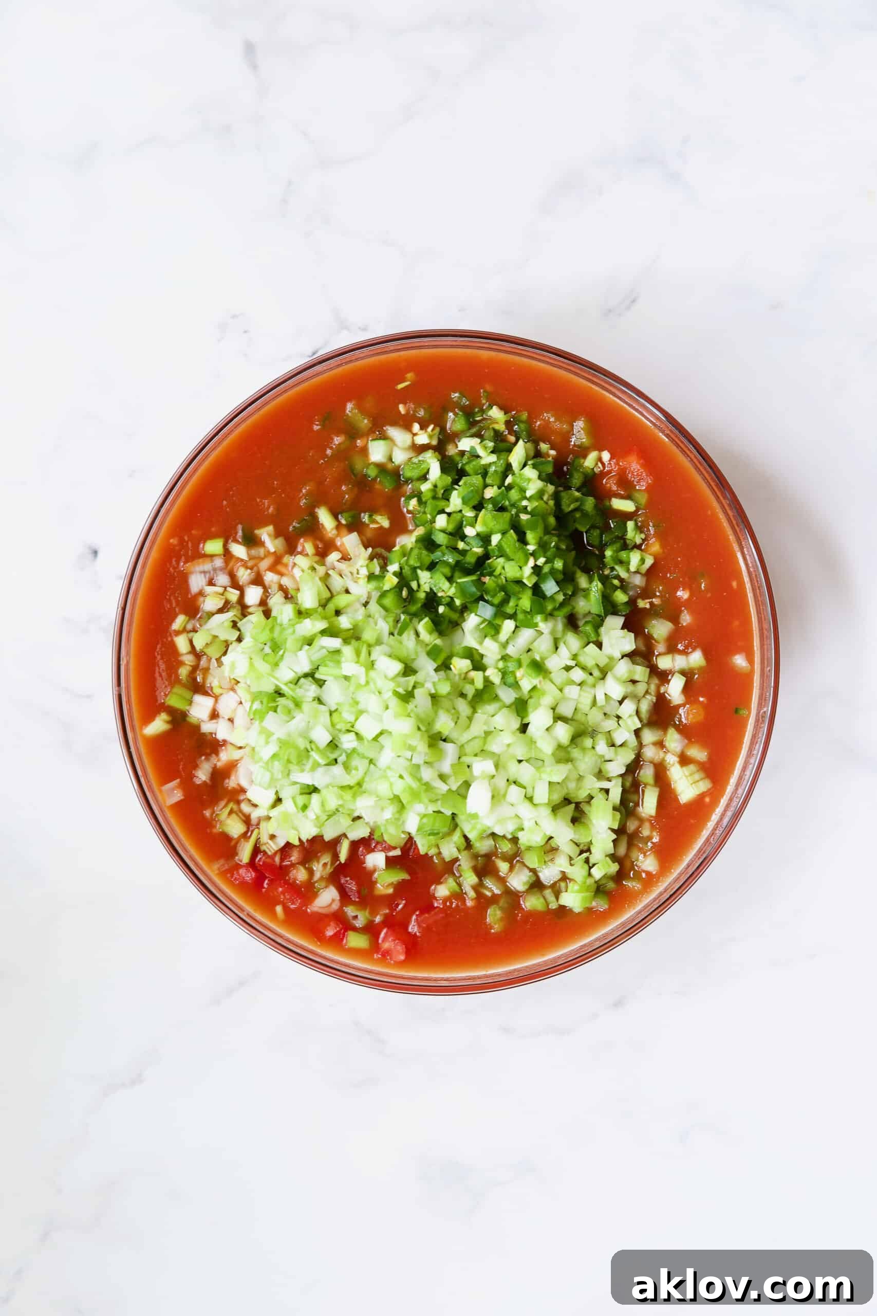 Finely chopped vegetables being added to the tomato juice base in a large glass bowl, illustrating the preparation of Very Veggie Gazpacho.