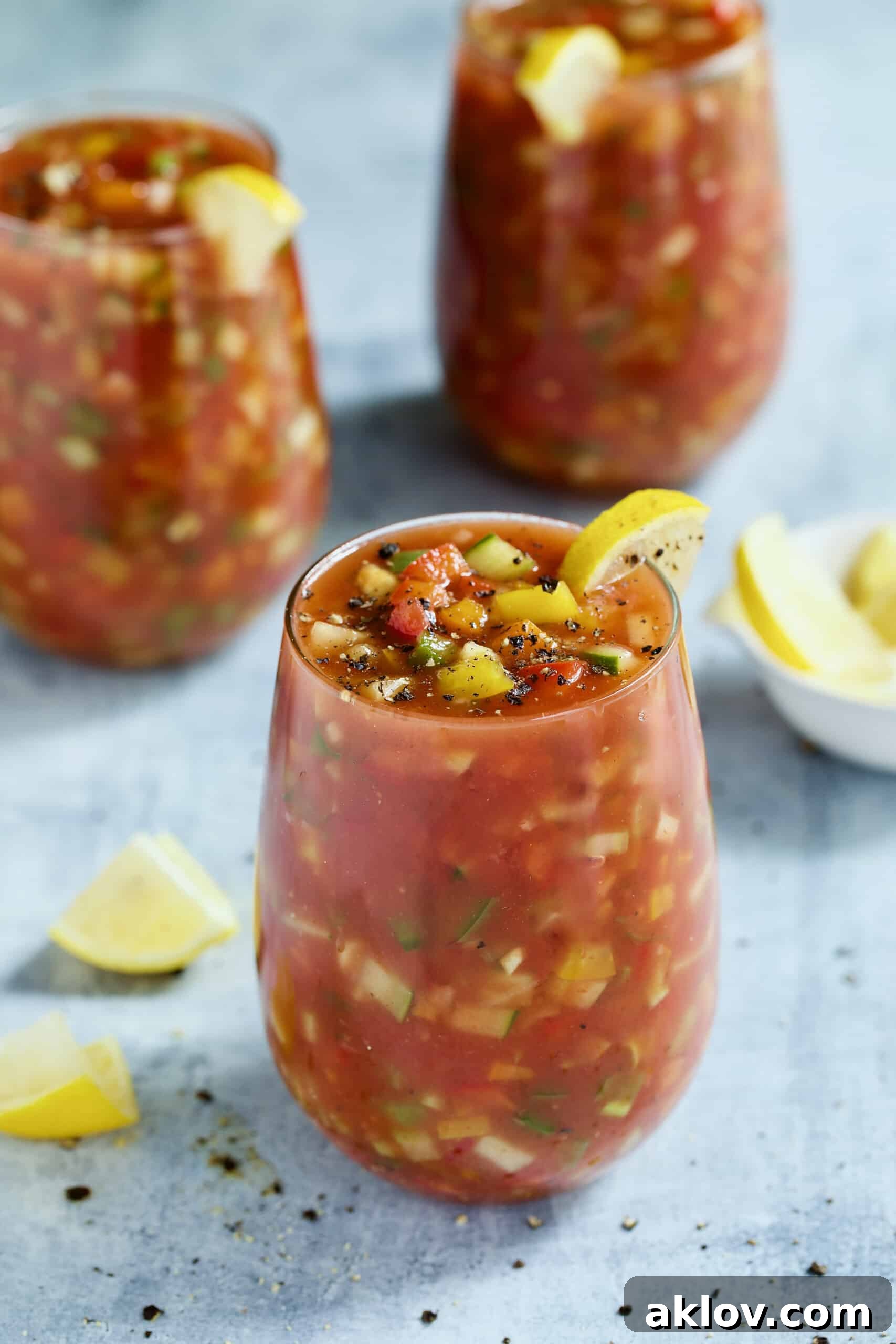 Three elegant glasses of Very Veggie Gazpacho garnished with fresh lemon wedges, presented on a light blue background for a refreshing summer meal.