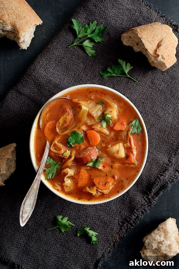 A single serving of Old-Fashioned Potato Cabbage and Kielbasa Soup in a white bowl, elegantly presented with a gray napkin and a slice of bread.