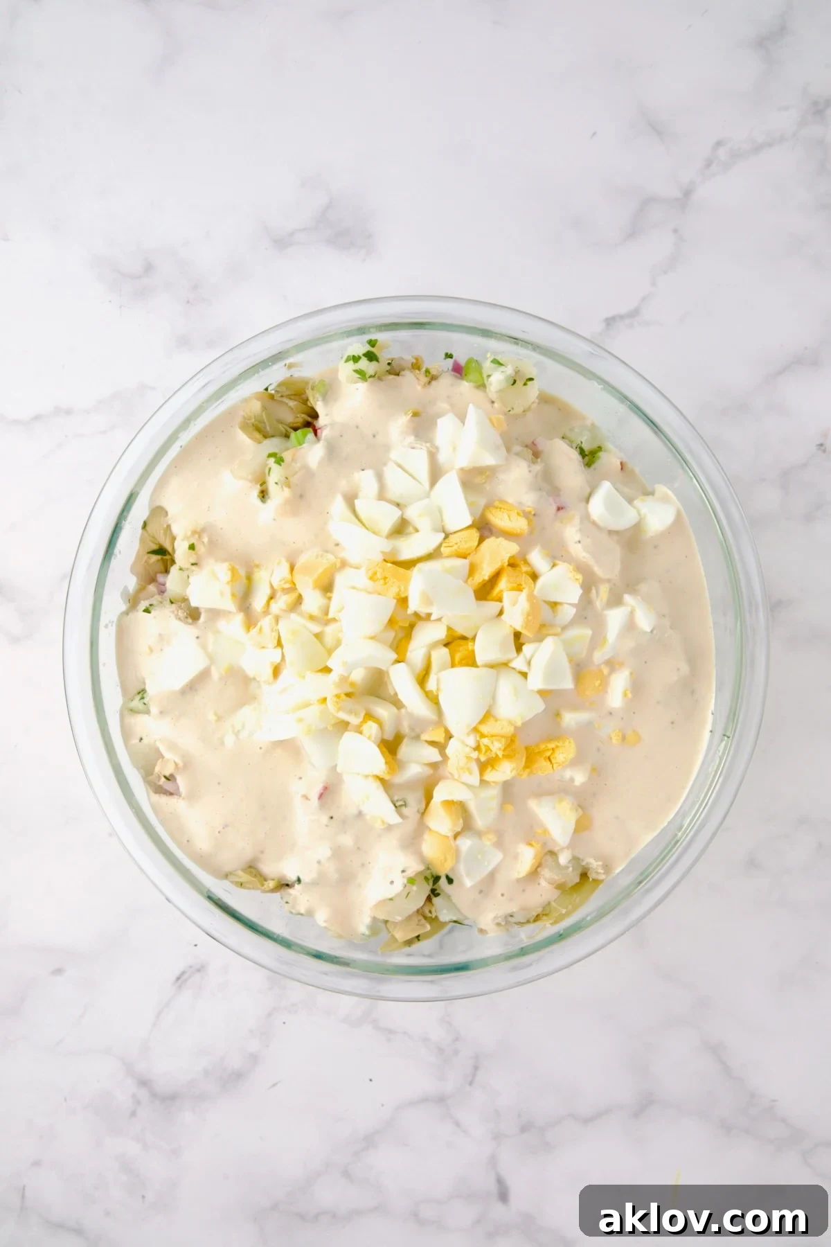 The creamy horseradish dressing being poured generously over the potato and vegetable mixture, with chopped hard-boiled eggs already added, in a large glass bowl, ready for final mixing.