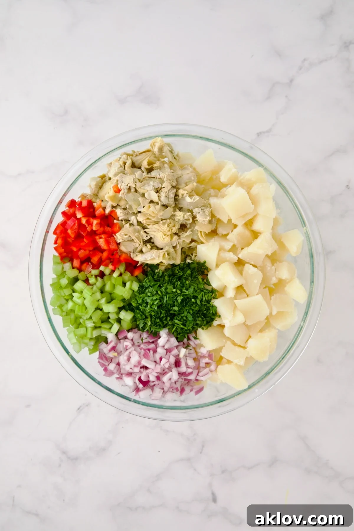 All the salad ingredients, including cooled potatoes, vibrant red onion, crisp celery, colorful red bell pepper, earthy artichoke hearts, and fresh parsley, waiting to be mixed in a large clear glass bowl.