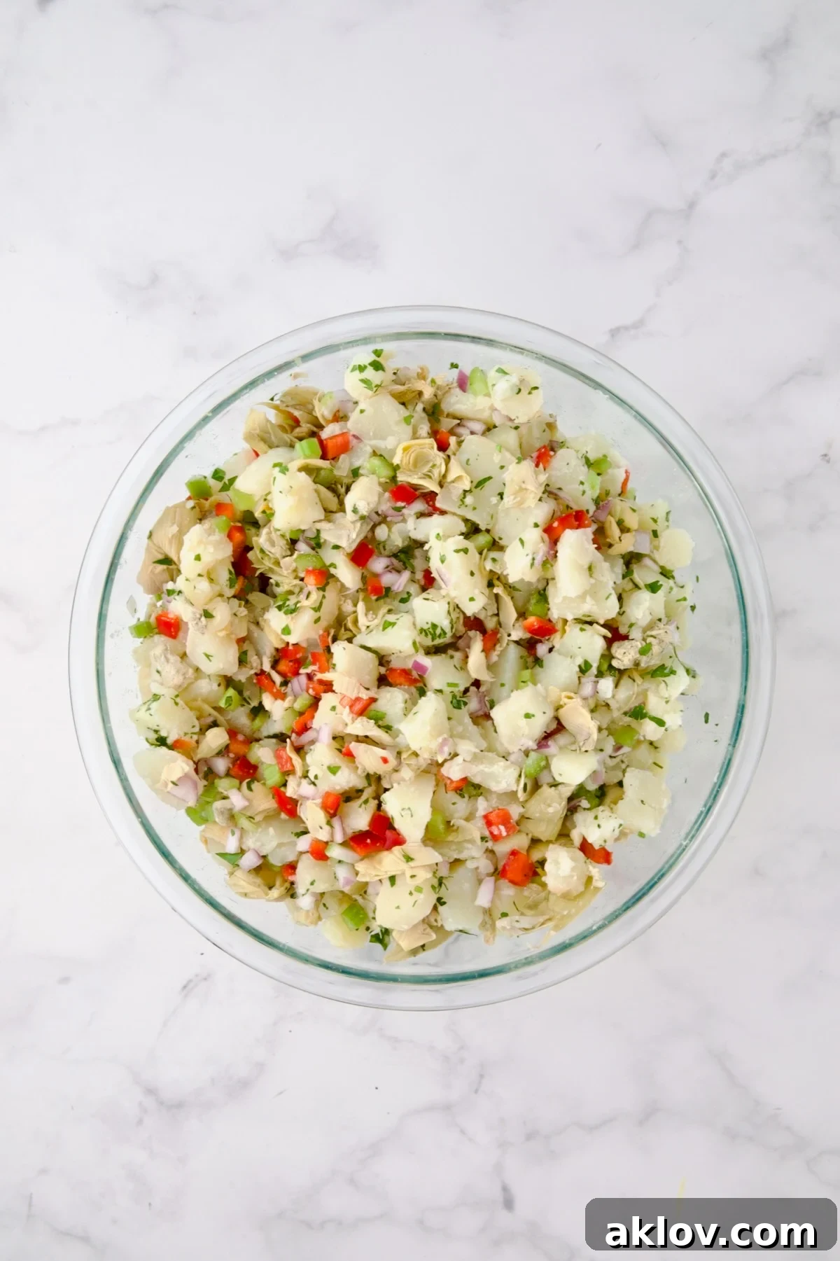 The mixed salad ingredients (potatoes, red onion, celery, red bell pepper, artichoke hearts, and parsley) in a large glass bowl, gently tossed together to ensure even distribution without mashing.