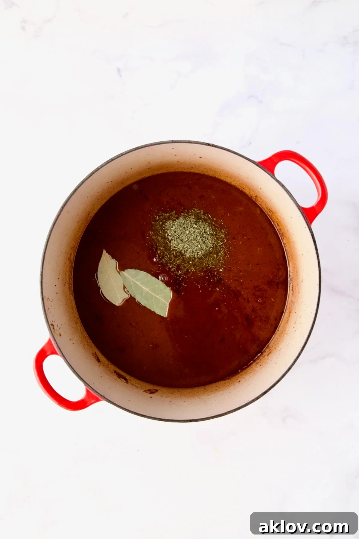Dried herbs and broth added to the short ribs in a red Dutch oven, preparing for slow braising.