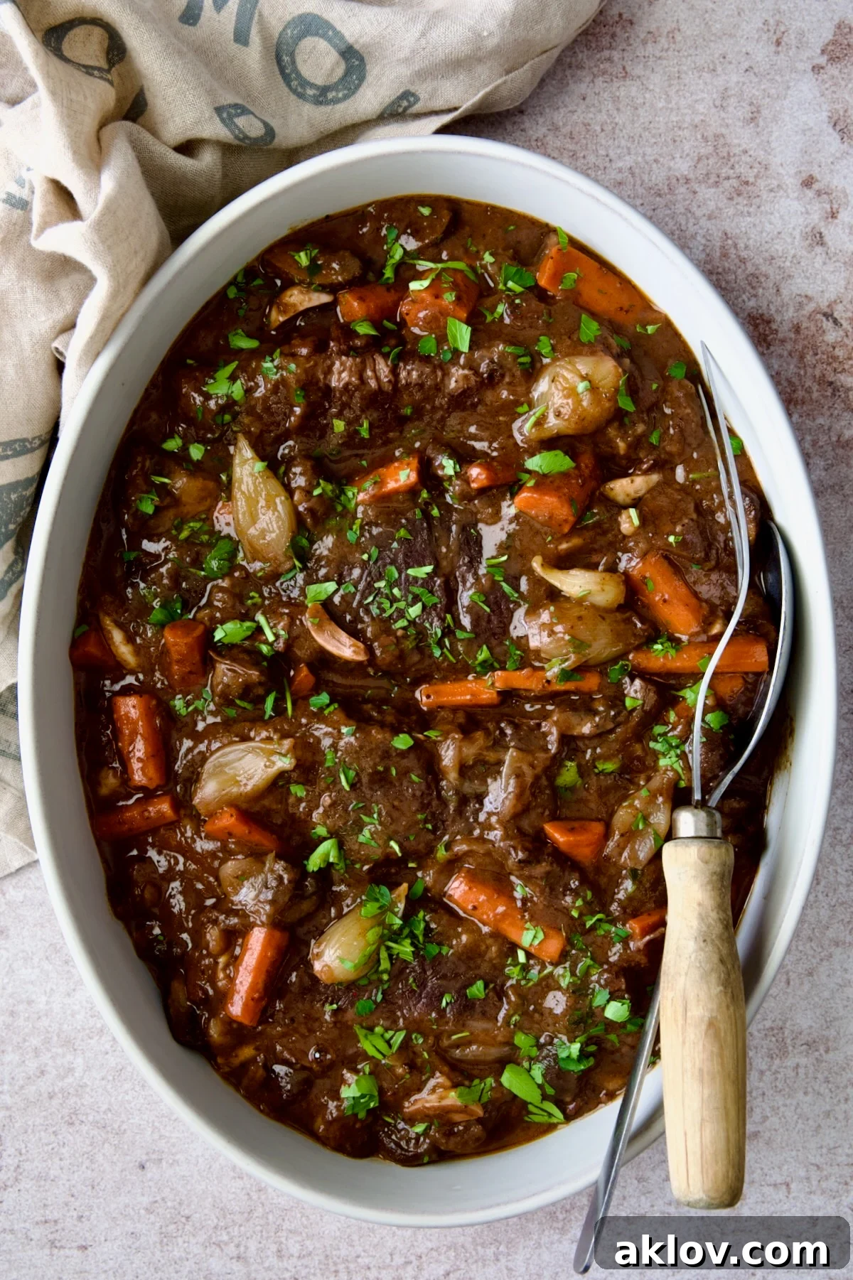 A stunning presentation of Short Ribs Bourguignon in a white oval serving dish with meat fork and serving spoon.