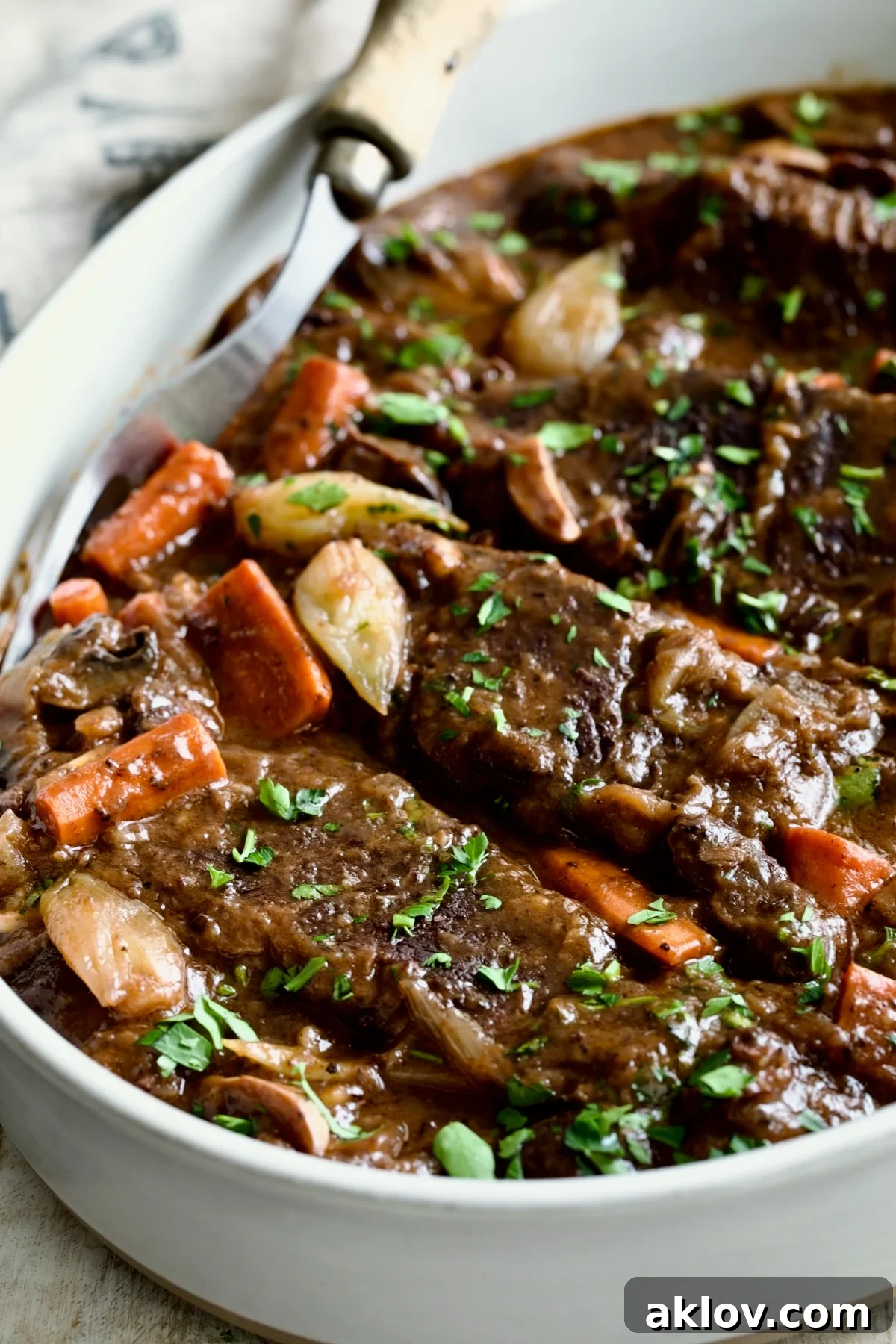 A generous serving of Short Ribs Bourguignon in a white oval serving bowl with a meat fork, ready for plating.