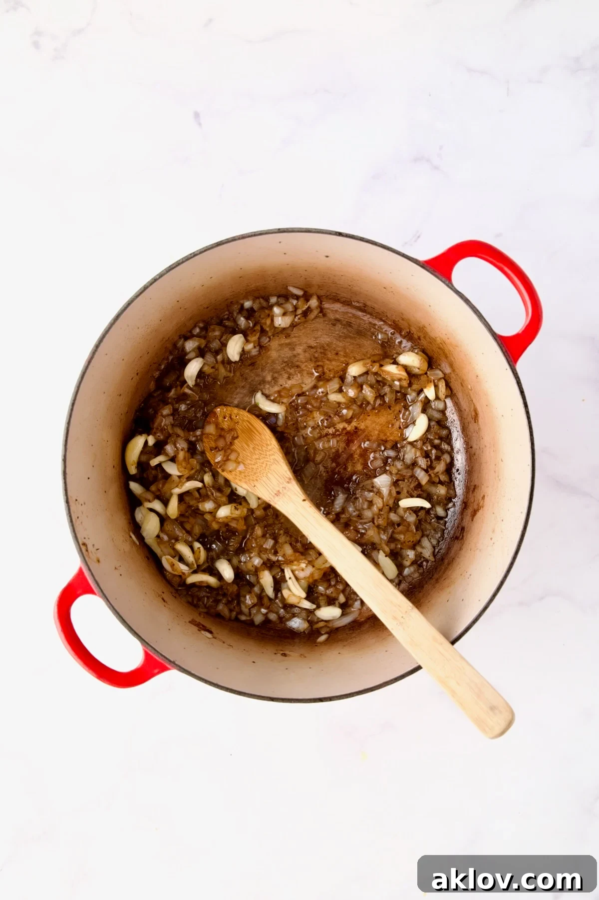 Onions and sliced garlic being stirred together with a wooden spoon in a red Dutch oven, releasing their aromas.