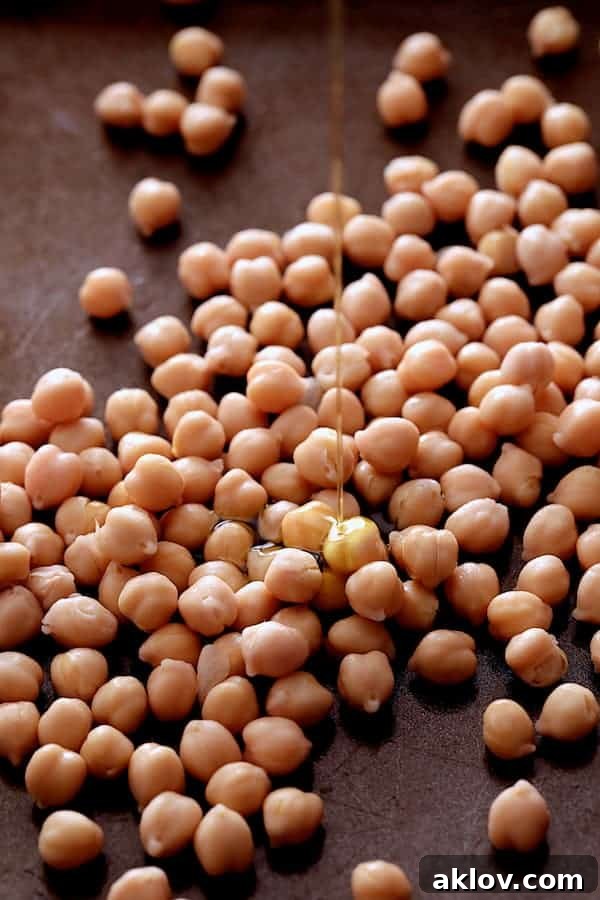 Close-up photo of chickpeas on a baking sheet, being drizzled with olive oil and seasoned with salt and pepper before roasting.