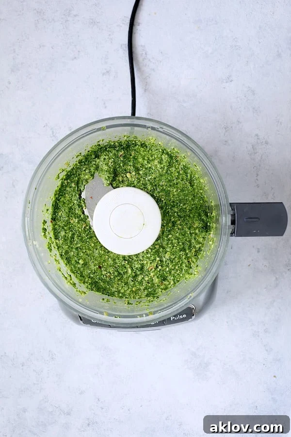A vibrant, emerald green Spinach Pesto Sauce, freshly blended to a smooth consistency, rests in the basin of a food processor, signaling its readiness for the next stage of culinary creation.