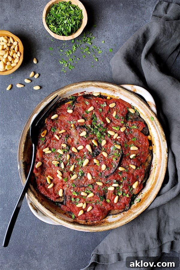 Photo of Healthier Turkish Eggplant Casserole - Imam Bayildi on dark gray background garnished with chopped parsley and pine nuts.