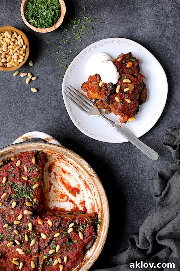 Photo of one piece of Turkish Eggplant Casserole - Imam Bayildi on a white plate with a dollop of yogurt the rest of the dish with wedge removed. A single, perfectly portioned slice of Turkish Eggplant Casserole (Imam Bayildi) is presented on a white plate, elegantly accompanied by a creamy dollop of yogurt, with the remaining casserole visible in the background.