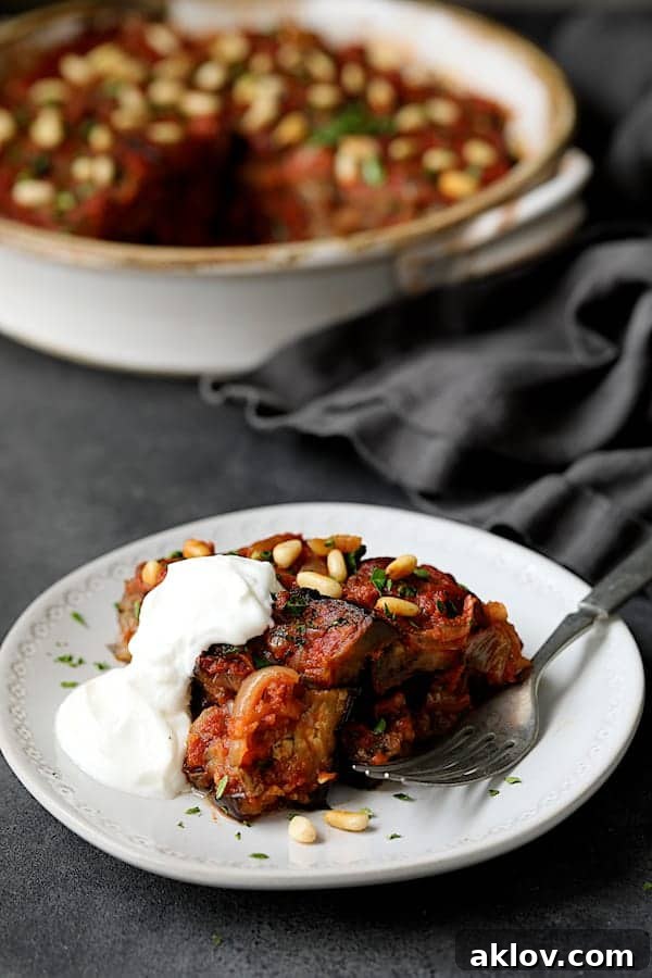 Close-up photo of one serving of Turkish Eggplant Casserole - Imam Bayildi on white plate with a dollop of yogurt with rest of the dish in the background.