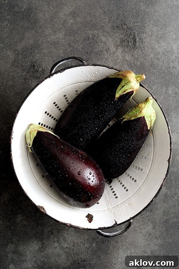 Photo of three freshly washed eggplant in aged white colander. Three vibrant purple eggplants rest gently in a rustic white colander, ready for preparation.