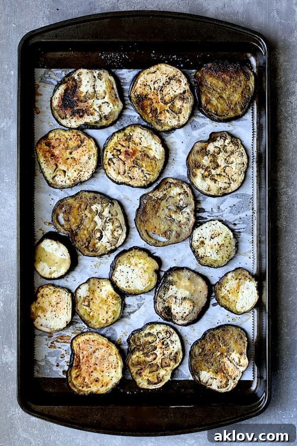 Photo of baked eggplant on parchment paper on baking sheet. Perfectly baked eggplant slices, lightly browned and tender, are neatly arranged on parchment paper on a baking sheet, showcasing their ready state for layering.