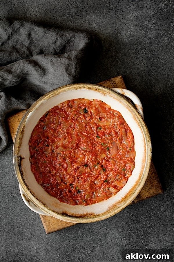 Photo of first layer of tomato sauce in round clay baking dish. A generous layer of rich tomato and onion sauce covers the bottom of a round clay baking dish, forming a vibrant base for the casserole.