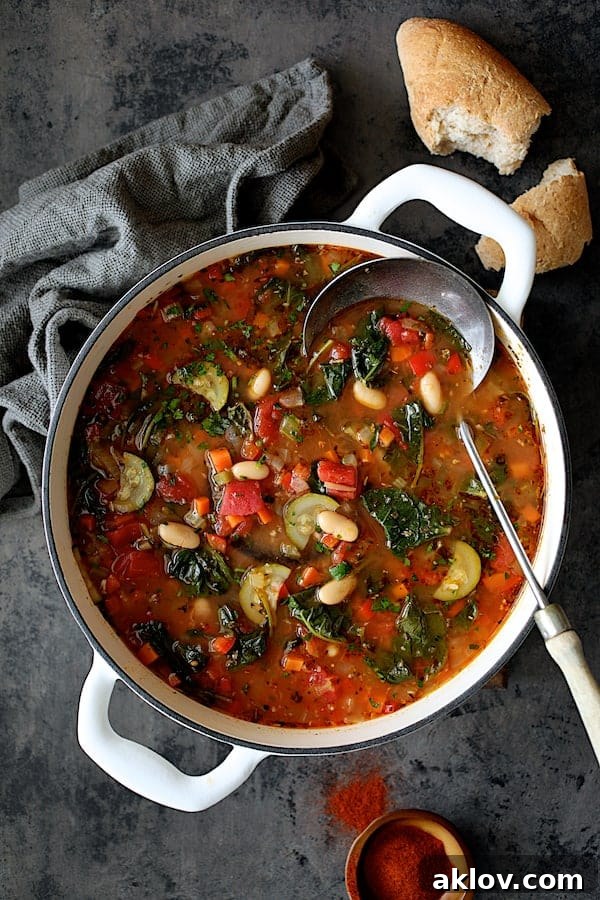 Photo of Smoky Spanish Vegetable and White Bean Soup with Kale in white Dutch oven with bread and paprika spilled on surface.