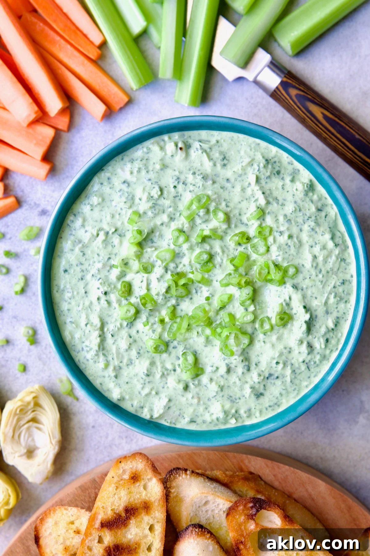 Spinach Artichoke Dip in round blue bowl garnished with scallions and carrot and celery sticks and bread round the bowl.