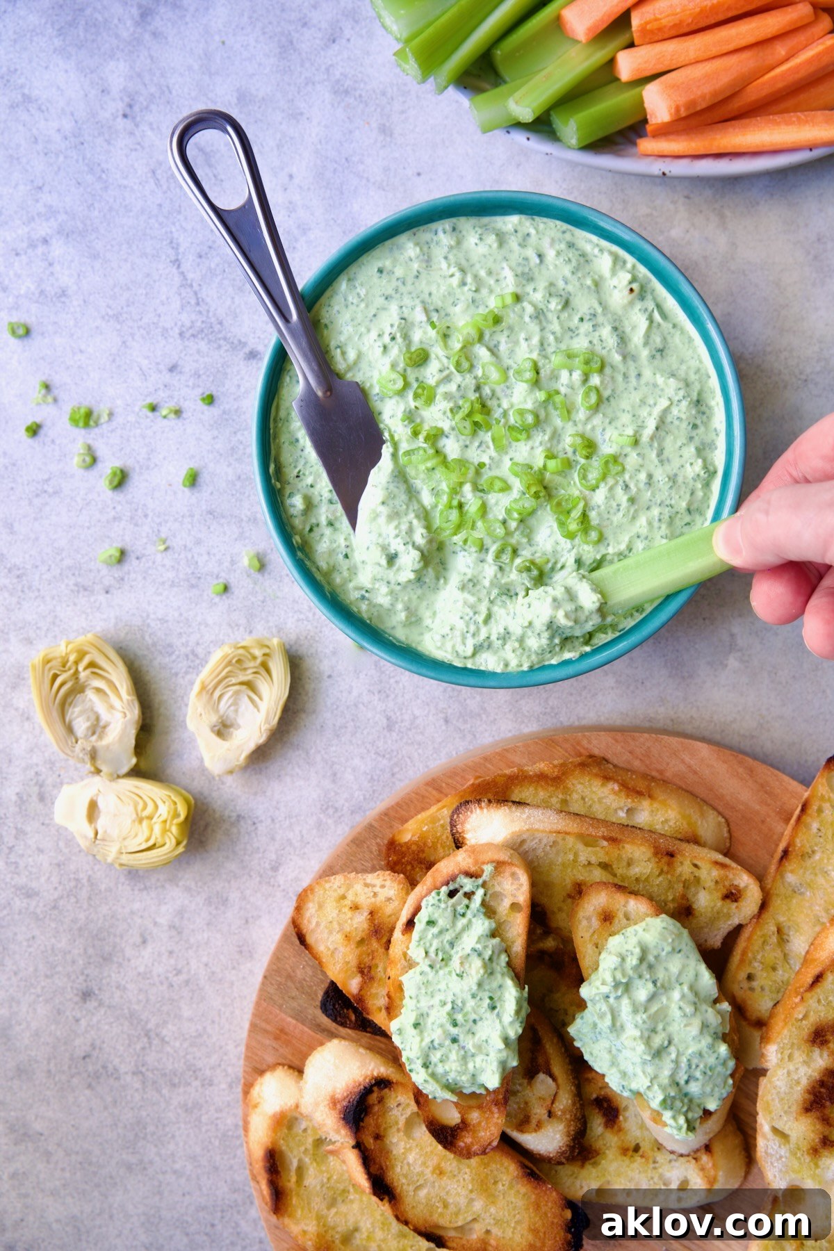 Spinach Artichoke Dip in round blue bowl garnished with scallions and carrot and celery sticks and bread round the bowl and person dipping celery stick into dip,.