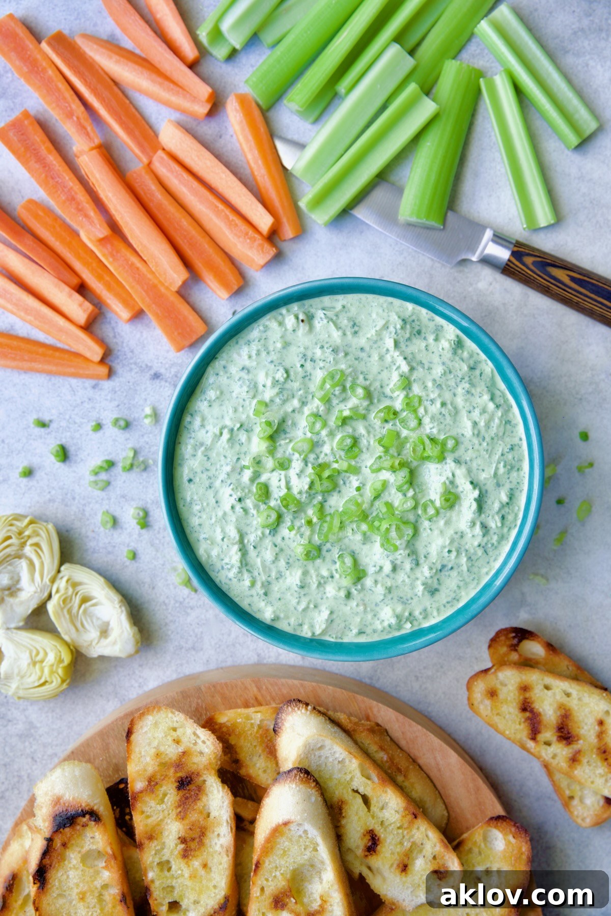 Spinach Artichoke Dip in round blue bowl garnished with scallions and carrot and celery sticks and bread round the bowl.