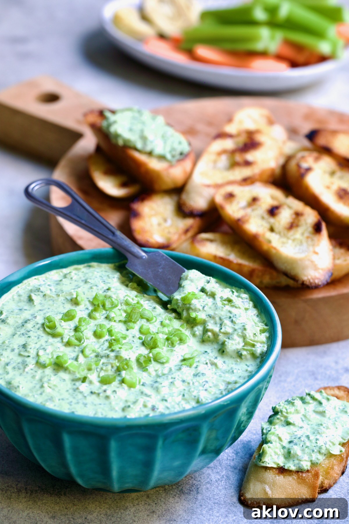 Spinach Artichoke Dip in round blue bowl garnished with scallions and carrot and celery sticks and bread round the bowl.