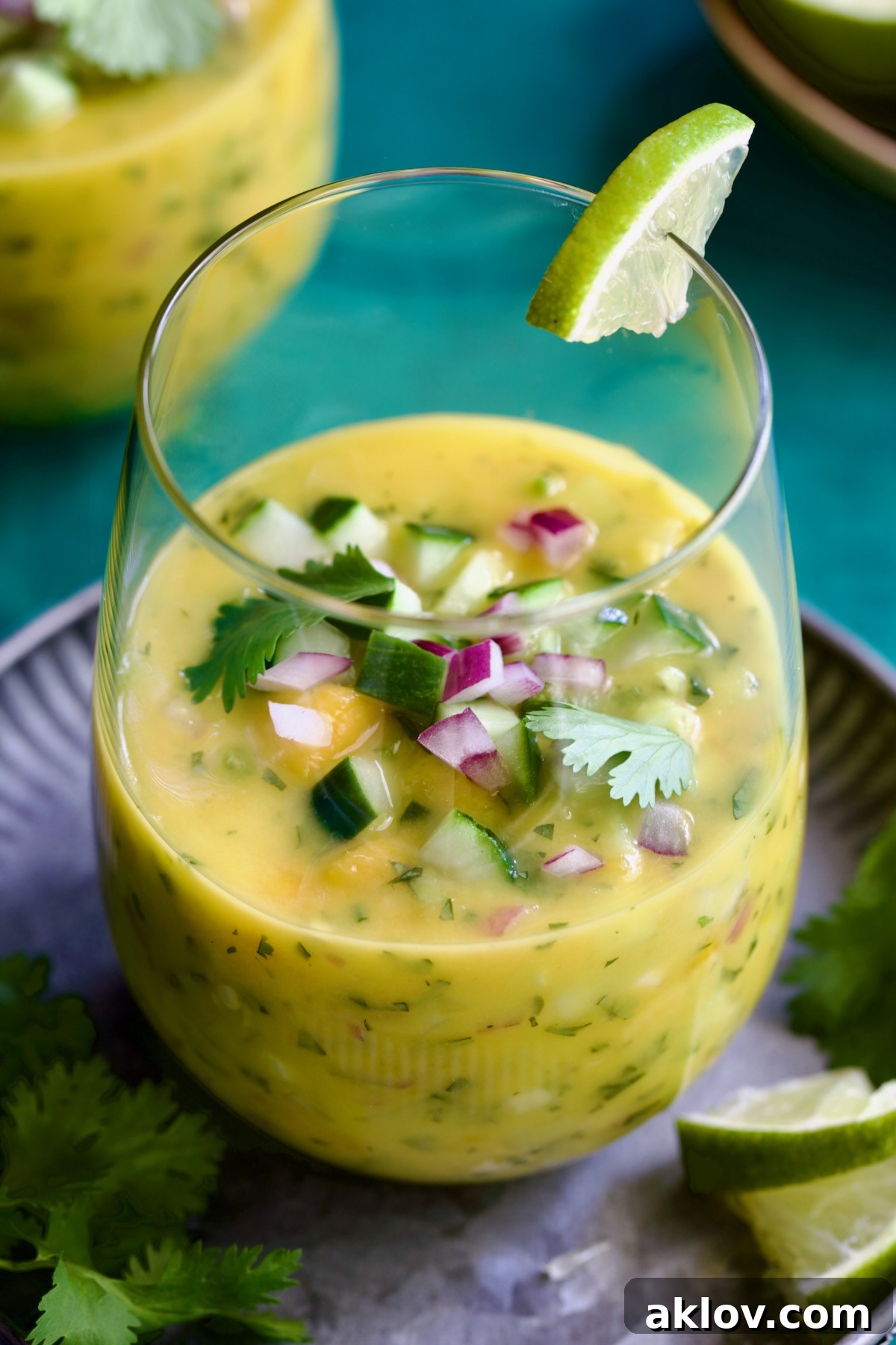 A single glass of luminous Mango Gazpacho rests on a sleek metal tray, elegantly adorned with a sprig of cilantro and a vibrant lime wedge, ready to be enjoyed.