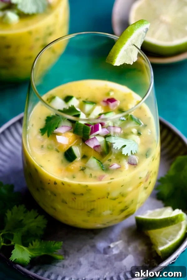 Vibrant Mango Gazpacho in a clear glass on a rustic metal tray, garnished with fresh cilantro and bright lime wedges. Perfect for a hot summer day.