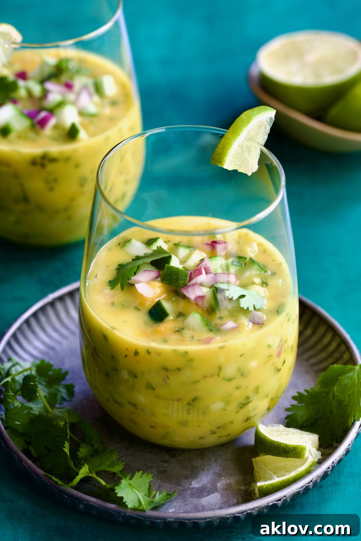 Two glasses of Mango Gazpacho with one on metal tray garnished with cilantro and lime wedges.