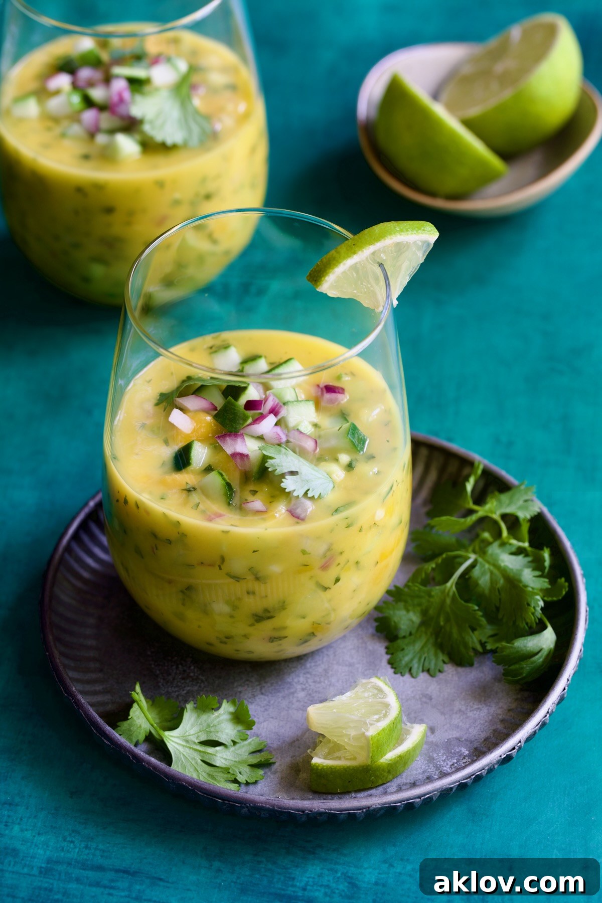 Two glasses of Mango Gazpacho with one on metal tray garnished with cilantro and lime wedges.
