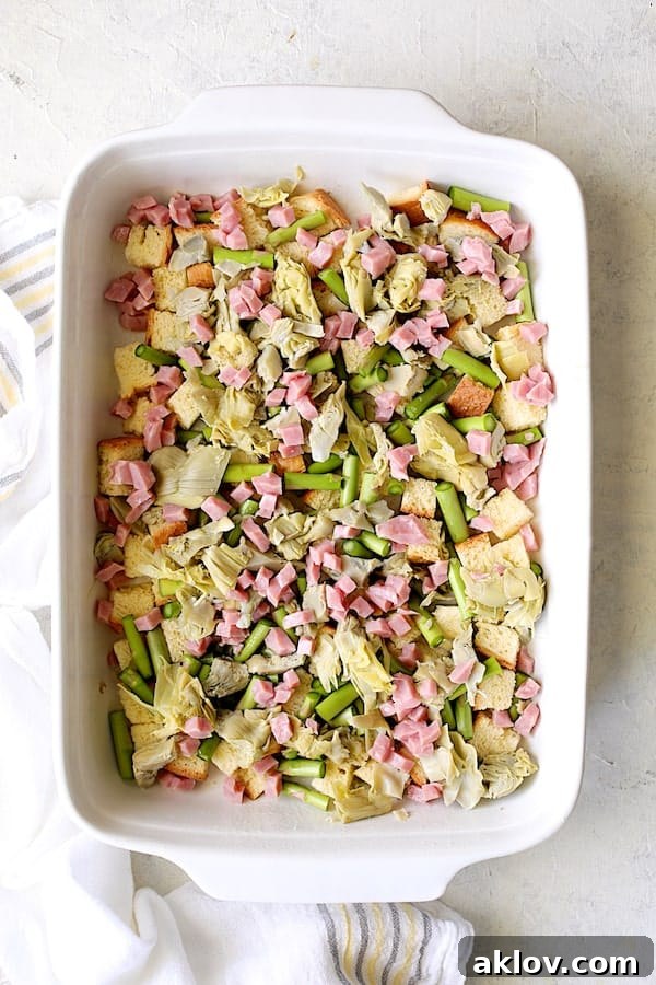 Layers of brioche, fresh asparagus stems, chopped artichokes, and diced ham in a white baking dish.