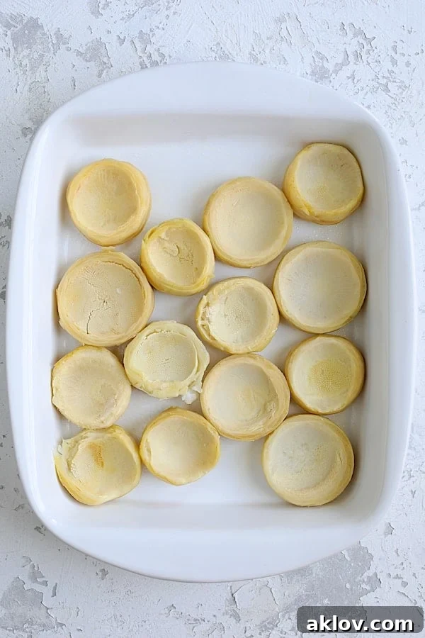 Fourteen empty, perfectly trimmed artichoke bottoms neatly arranged in a white baking dish, ready for filling.
