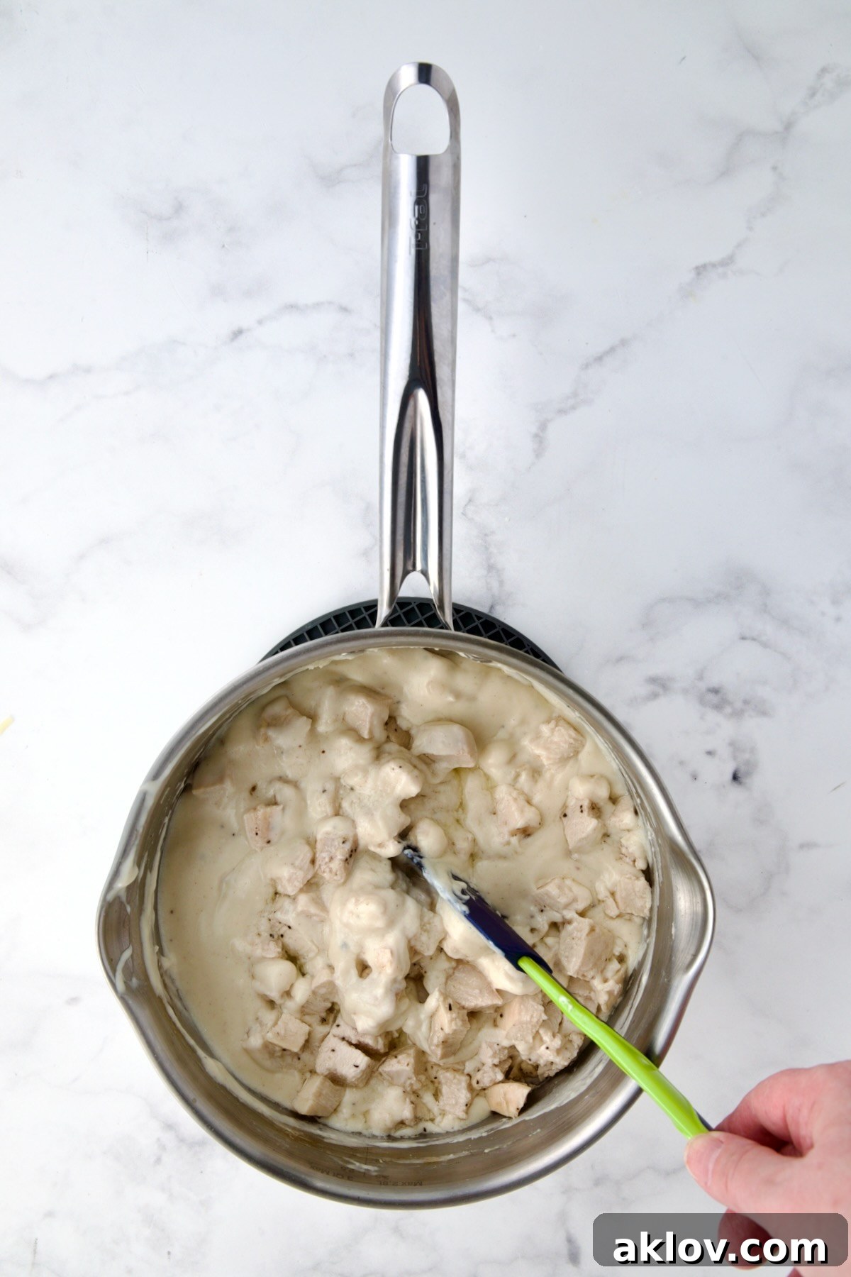 Cooked chicken being gently stirred into the rich, creamy cheese sauce with a spatula, ensuring even coating.