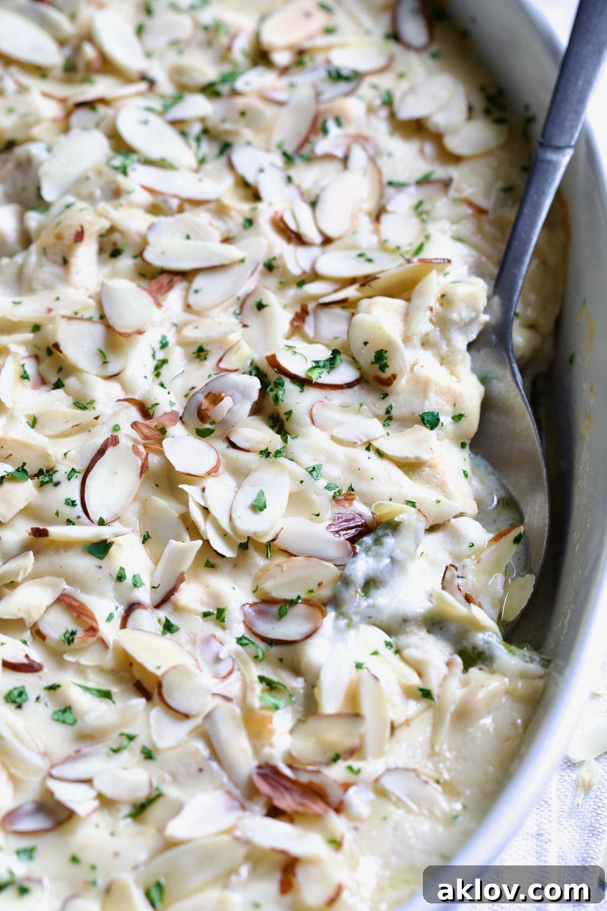 A serving spoon lifting a portion of creamy Chicken Divan with Asparagus, adorned with sliced almonds, from a baking dish, showcasing its delicious layers.