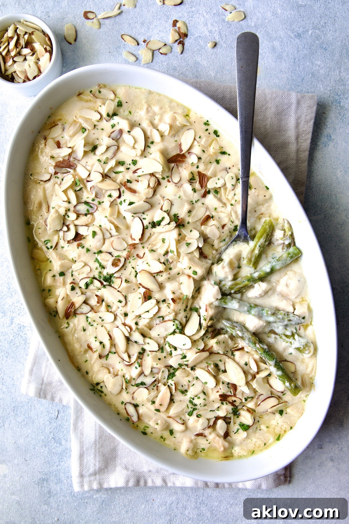 A serving spoon revealing layers of tender asparagus and succulent chicken in a creamy Chicken Divan, garnished with sliced almonds, in an oval baking dish.