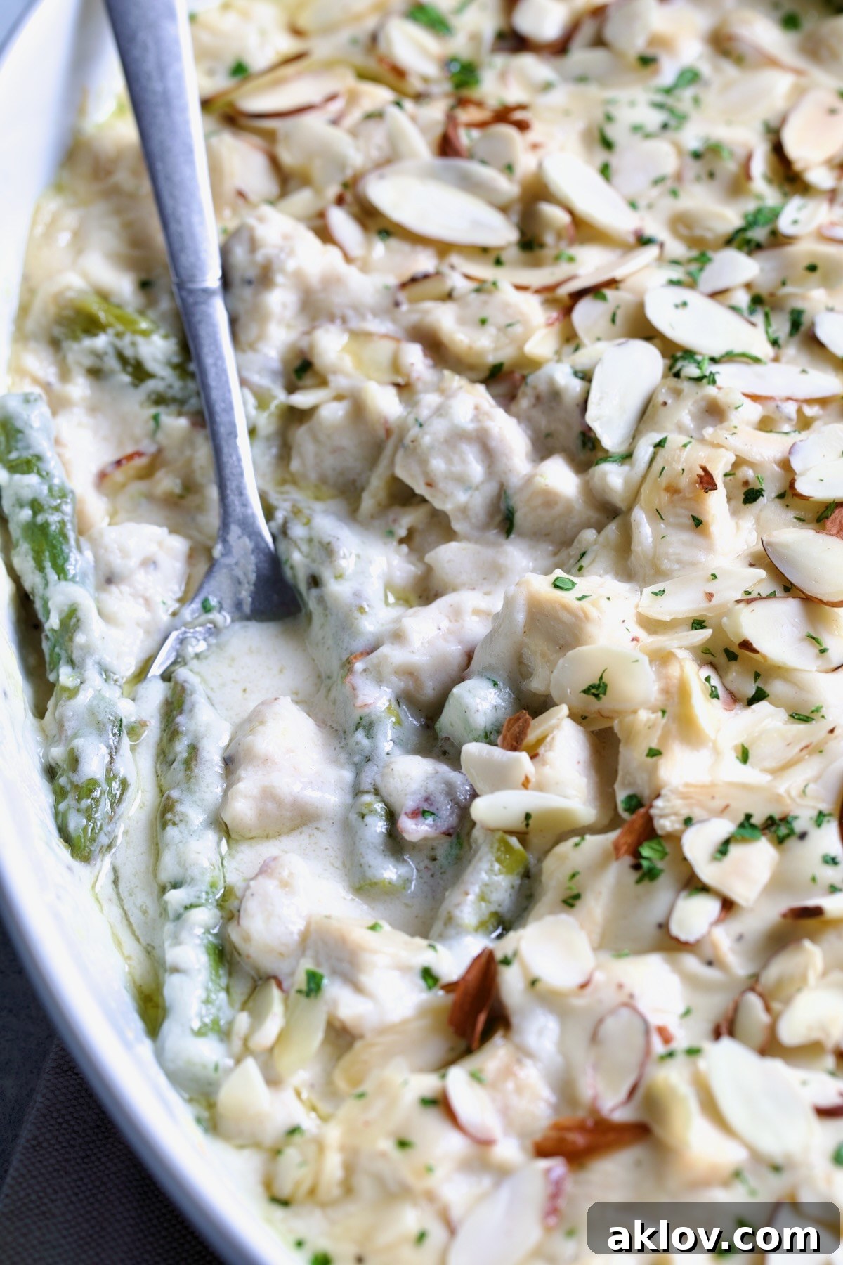 Close-up of a serving spoon scooping creamy Chicken Divan with Asparagus, garnished with almonds, from an oval baking dish.