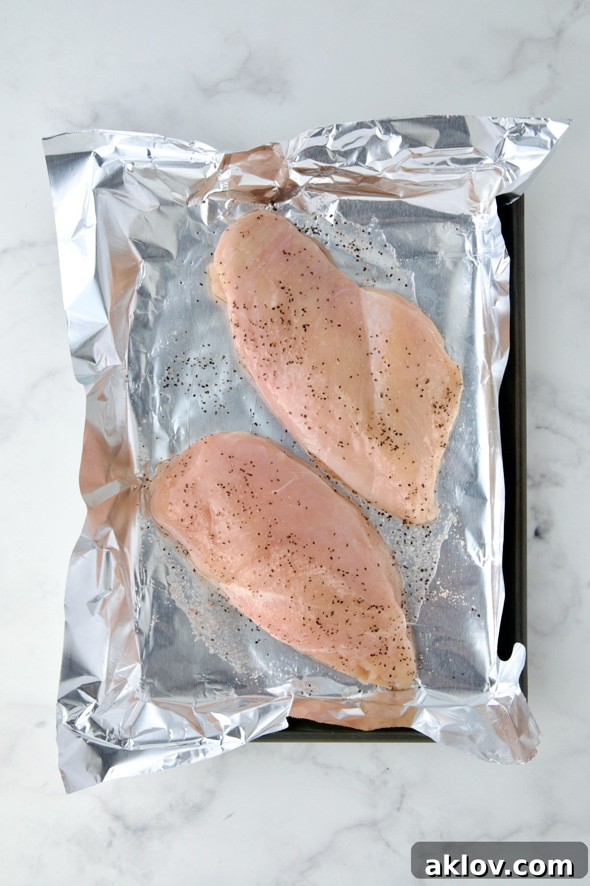 Two raw chicken breasts seasoned with salt and pepper on an aluminum foil-lined baking sheet, ready for roasting.