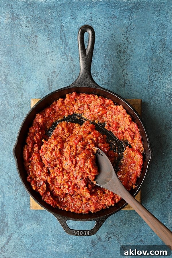 A wooden spoon stirs rich, red lentil and tomato sauce in a cast iron skillet, showcasing the deep red color and comforting texture of the simmering base.