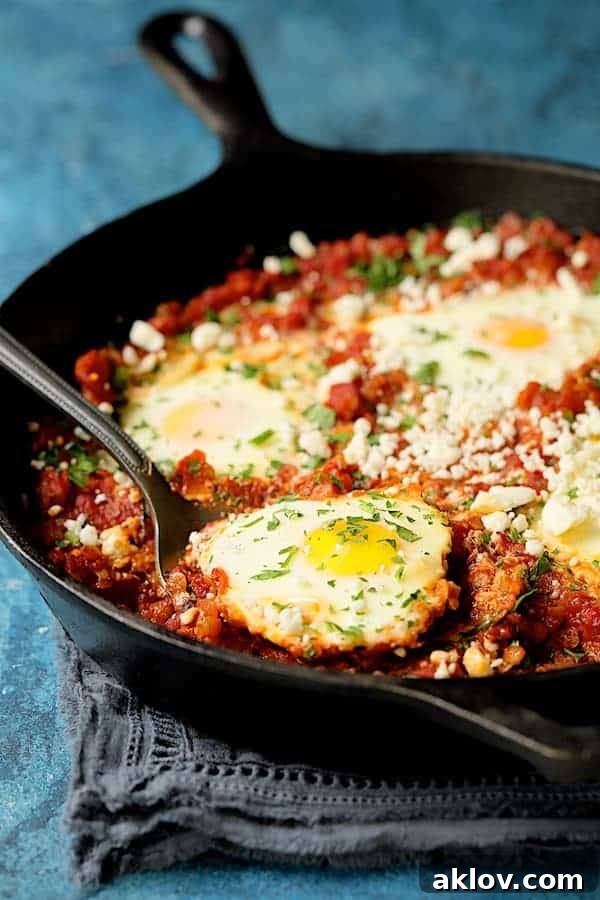 A spoon lifts a perfectly poached egg out of the bubbling Shakshuka with Red Lentils and Feta Cheese in a cast iron skillet.