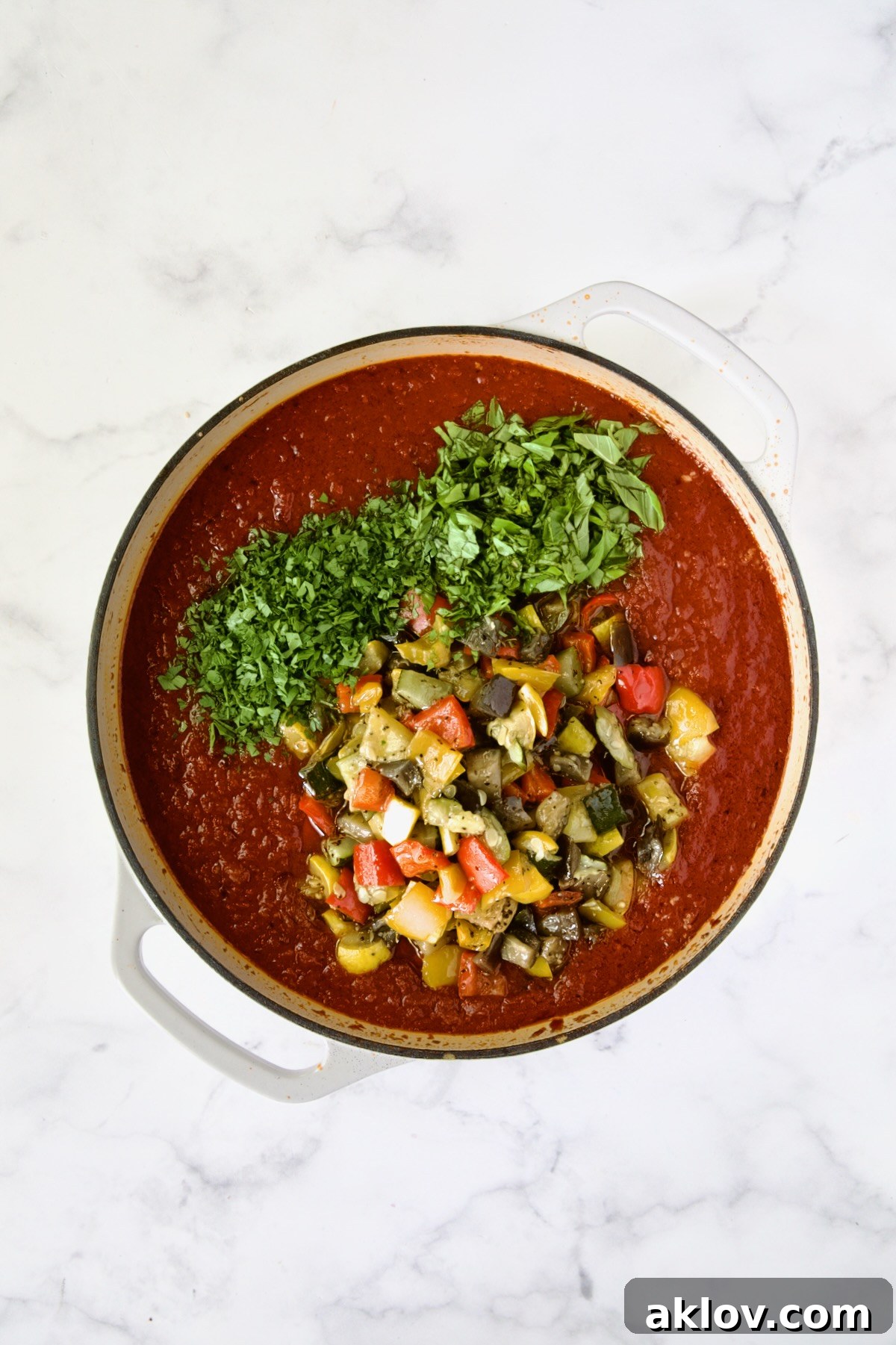 Roasted vegetables, parsley and fresh basil added to tomato sauce in white enamel cast iron skillet.