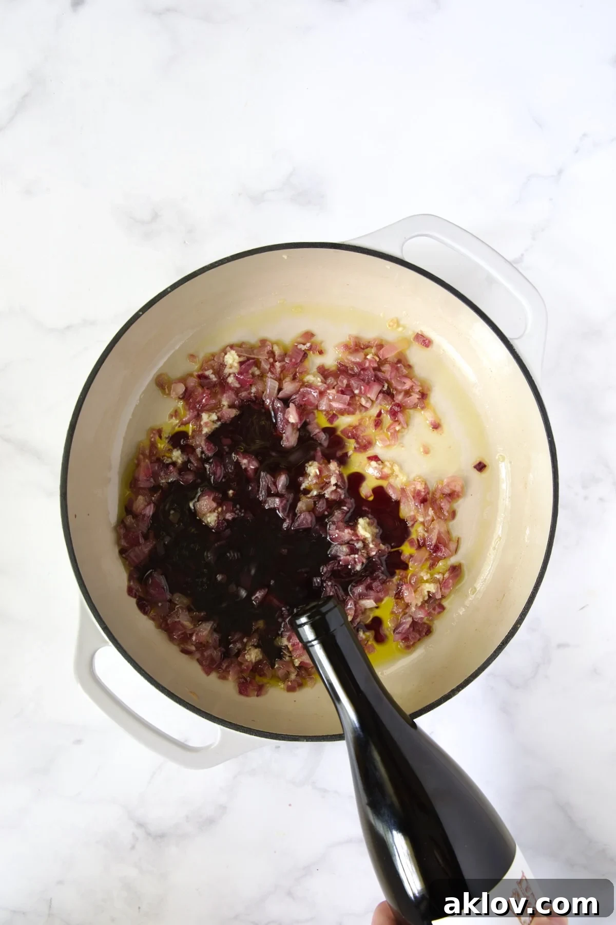 Red wine being poured onto onions from bottle into white enamel cast iron skillet.