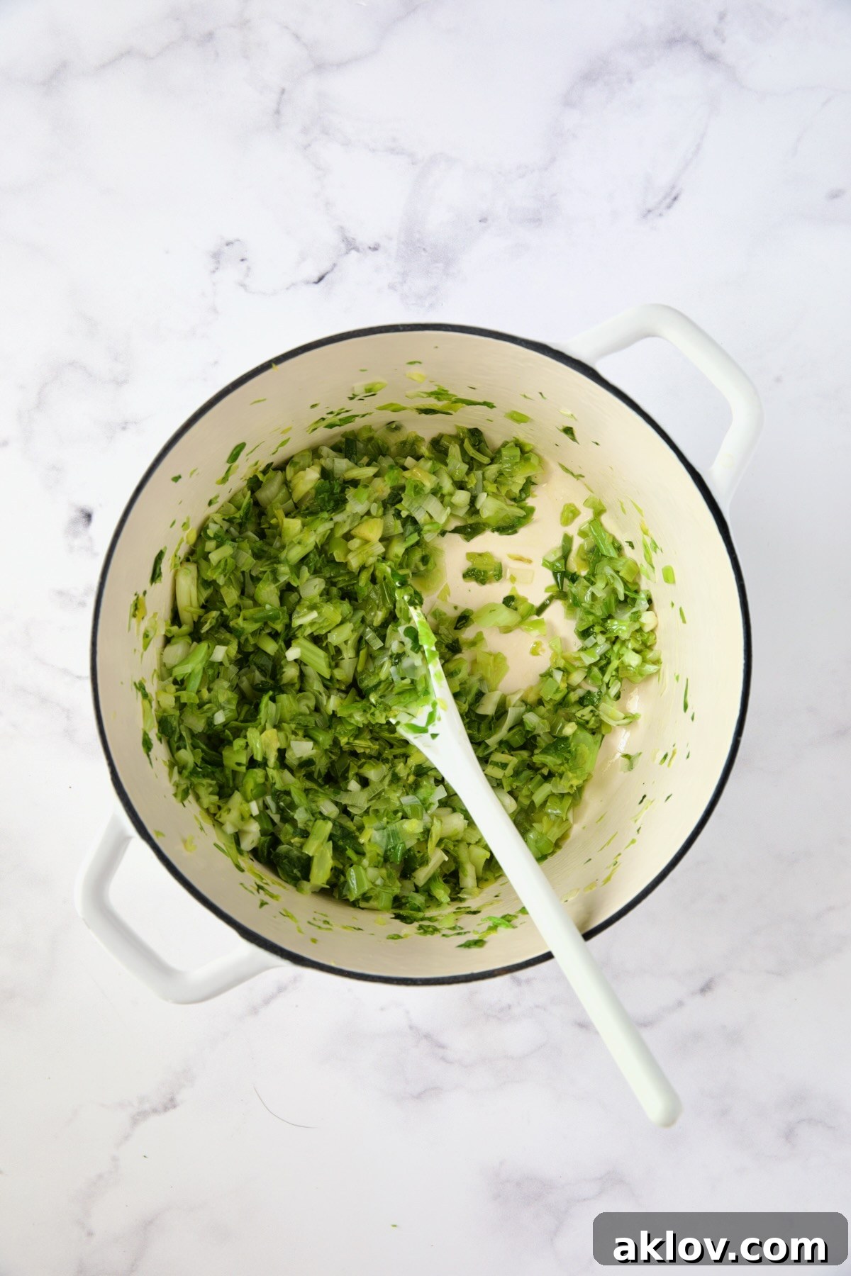 Scallions and celery leaves being sautéed together in a white Dutch oven, stirred with a white mixing spoon.
