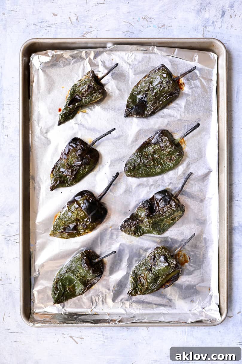 Photo of perfectly roasted Poblano peppers, their skins charred and softened, resting on a foil-lined baking sheet after steaming.