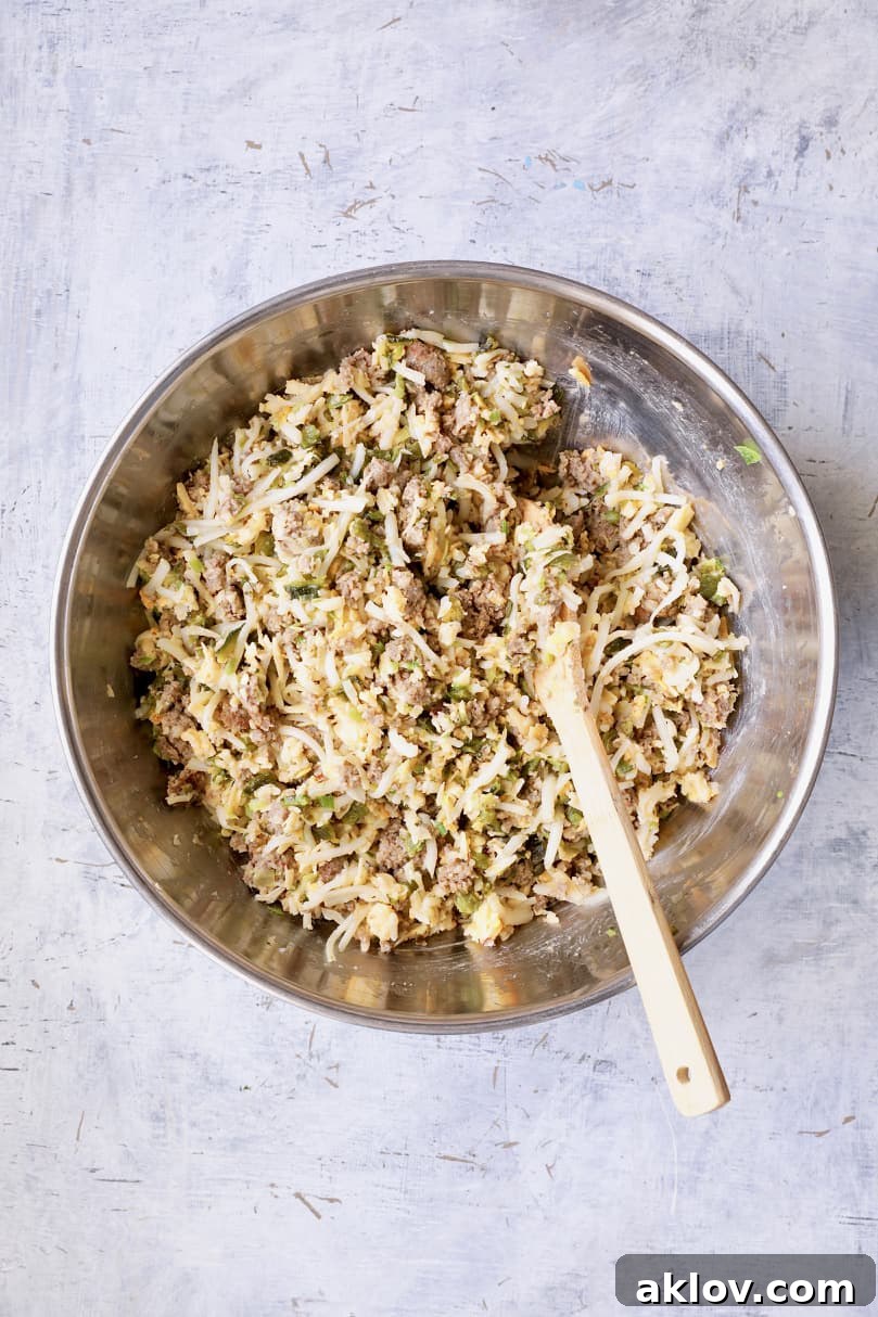Photo of all the savory ingredients for breakfast muffins—meat, potato, cheese, peppers—combined in a large stainless steel bowl, being stirred with a wooden spoon.