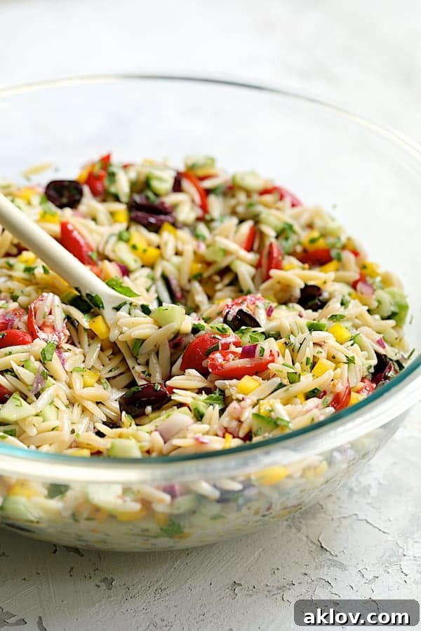 Greek Orzo Pasta Salad being gently mixed with a white spoon in a large glass bowl, showcasing the vibrant colors and uniform distribution of the ingredients before feta is added.