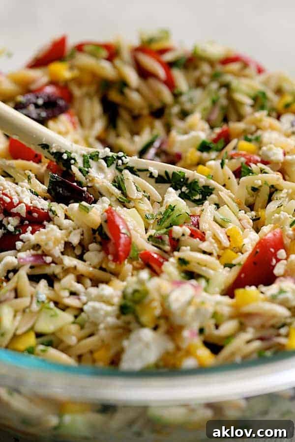 Greek Orzo Pasta Salad, already tossed with dressing, with crumbled feta cheese being gently added and mixed in, captured in a clear glass bowl on a white surface.