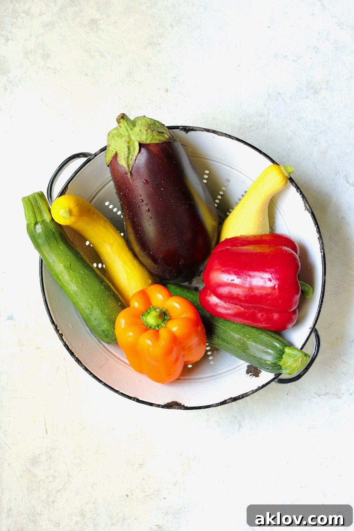 Freshly cut zucchini, yellow squash, eggplant, and bell peppers neatly arranged in a vintage white colander.