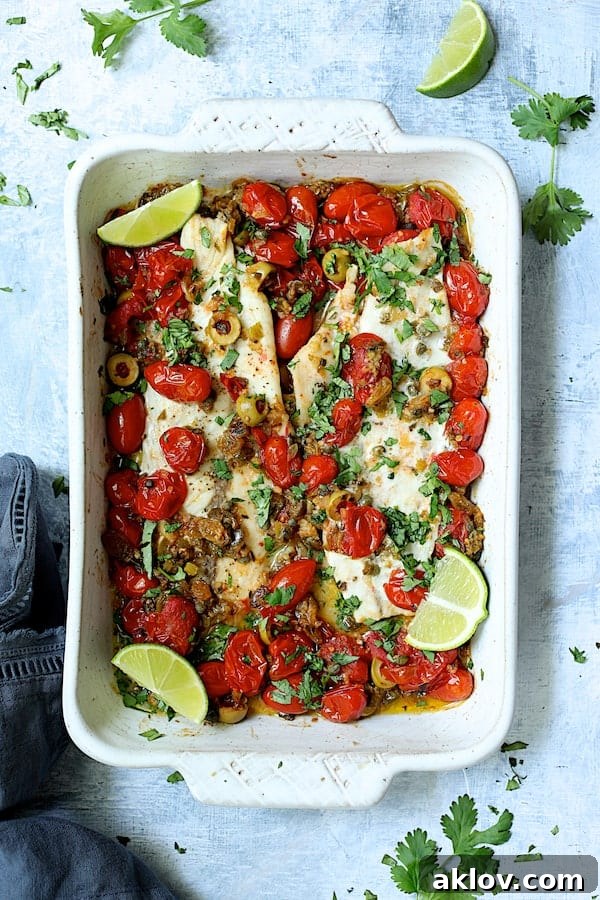 Photo of Baked Veracruz-Style Fish (Pescado A La Veracruzana) in white baking dish on light blue background garnished with lime wedges and cilantro.